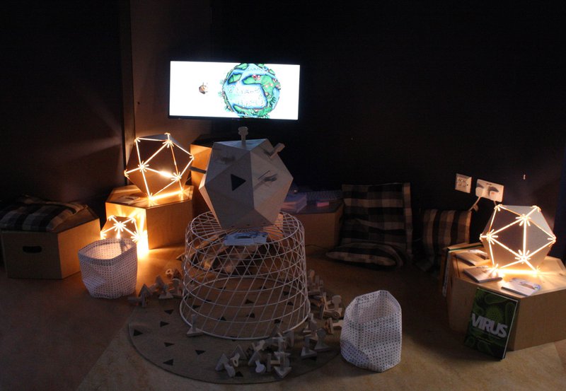 Viruscraft installation at the eden project. There is a wood virus in the centre, with a screen in the background with the game on it. There are lights in the shapes of viruses, and cushions.